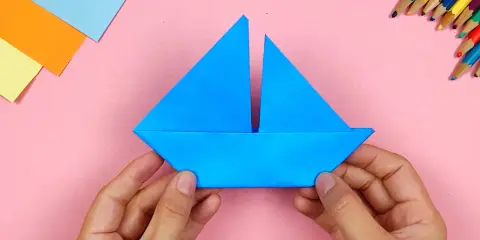 Hände falten ein blaues Papierboot. Bunte Stifte und Papier liegen bereit für weitere Bastelprojekte.