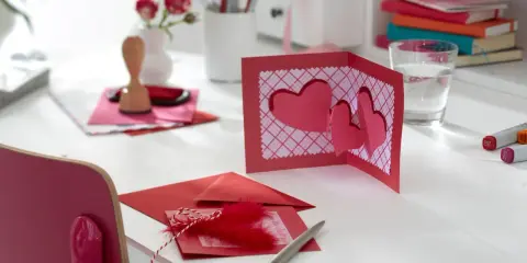 Eine 3D-Karte basteln zum Geburtstag: Rote Herzen auf einer Klappkarte, Stempel, Federn und Stifte liegen bereit.
