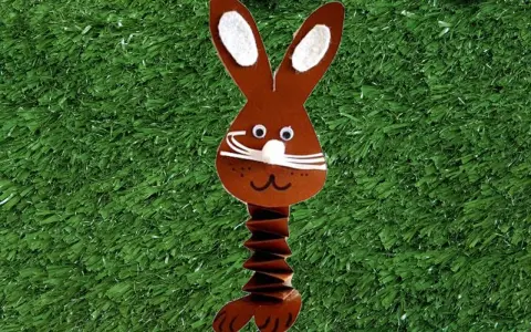 Brauner Papierhase mit Ziehharmonika-Körper, ein süßes Beispiel für Osterhasen basteln, auf grünem Kunstrasen mit bunten Eiern.