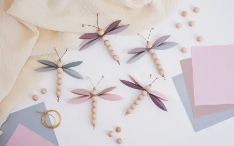 Vier selbstgebastelte Libellen aus Holzperlen und Papierflügeln, inspiriert von einer libelle basteln vorlage. Zarte Pastellfarben und ein Musselintuch schaffen eine frühlingshafte Atmosphäre.