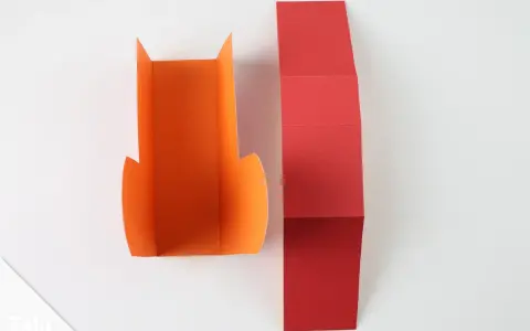 Zwei Bastelbögen, einer orange, der andere rot, liegen nebeneinander. Der rote Bogen ist gefaltet, bereit, um daraus Stiefel zu falten.