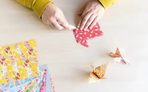 Hände falten Papier für eine Origami Kugel Anleitung. Mehrere bunte Origami-Papierbögen liegen bereit.