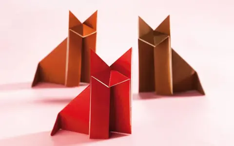 Drei origami Füchse, einer rot und zwei braun, sitzen auf einem rosa Untergrund.