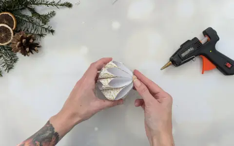 Hände basteln mit Klebepistole und Papier einen schönen DIY Christbaumschmuck. Tannenzweige und Orangenscheiben liegen bereit.