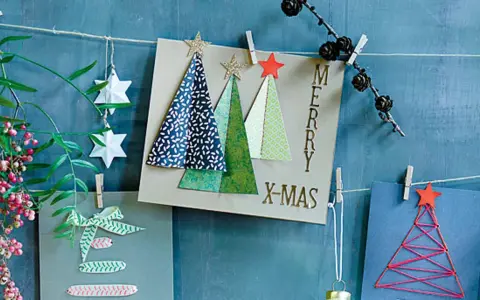 Selbstgebastelte Weihnachtskarten mit Bäumen und Sternen hängen an einer Schnur. Ein grüner Zweig mit Beeren und Papiersterne zieren die Szene.