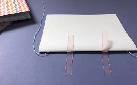 Beim Buch basteln werden weiße Blätter mit rosa Bändern und Faden zusammengefügt.