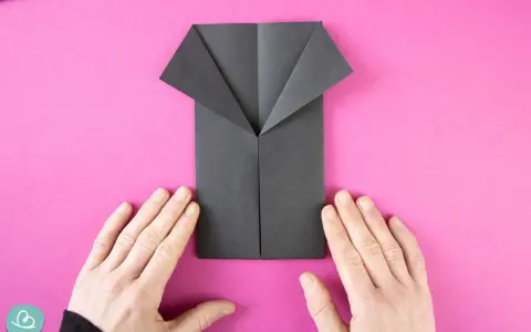 Ein schwarzes Origami-Hemd wird auf pinkem Hintergrund gefaltet. Hände halten das Papier.
