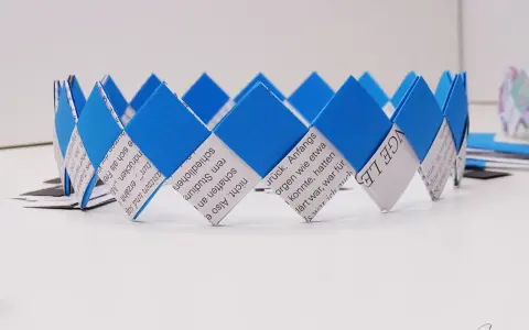 Eine blaue und weiße Papierkrone, die aus gefalteten Papierstreifen gefertigt wurde. Die Streifen sind mit Text bedruckt, was die Krone zu einem besonderen Kunstwerk macht.