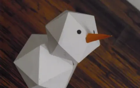 Geometrischer Schneemann, gefaltet aus Papier, mit orangefarbener Karotte als Nase und schwarzen Augen.