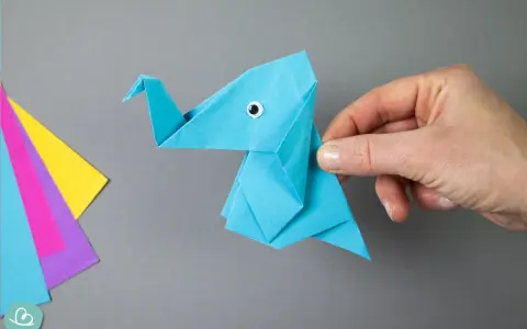 Ein blauer origami elefant mit einem kleinen Knopfauge wird von einer Hand gehalten. Daneben liegen bunte Papierbögen.