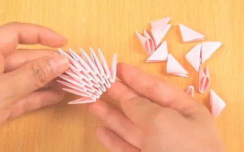 Hände bauen mit rosa Papierstücken ein 3D-Origami-Tier. Die Anleitung ist leicht verständlich.