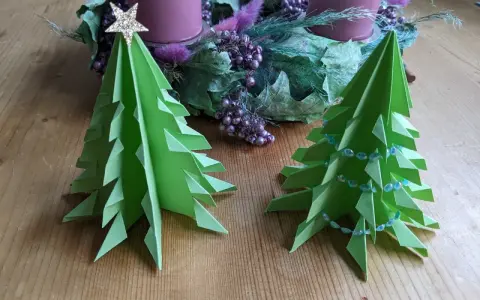 Zwei grüne, flache Origami Tannenbäume stehen auf einem Holztisch. Einer ist mit einer goldenen Sternspitze geschmückt, der andere mit einer Perlenkette. Im Hintergrund ein Adventskranz.