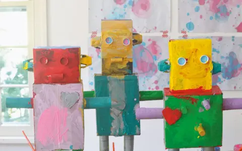 Drei bunte Roboter, die Kinder gebastelt haben, stehen nebeneinander. Sie sind aus Kartons und Rollen gefertigt und mit leuchtenden Farben bemalt.