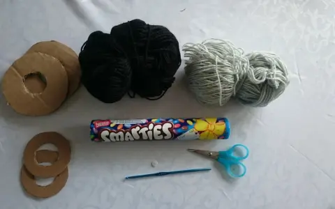 Materialien für ein Geldgeschenk: Pappringe, schwarzes und hellgraues Garn, Smarties-Rolle, Knopf, Häkelnadel und Schere. So lässt sich eine Hantel basteln.