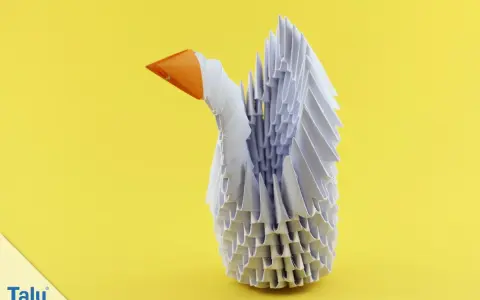 Ein weißer Schwan aus modularem Origami mit orangefarbenem Schnabel steht vor einem gelben Hintergrund.