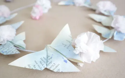 Zarte Origami-Schmetterlinge aus Buchseiten, verziert mit Tupfen und kleinen Papierblumen, bilden einen Kranz. Ein Hauch von origami frühling.