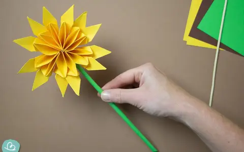 Eine Hand hält eine gelbe origami sonne mit einem grünen Stiel. Bastelmaterialien liegen daneben.