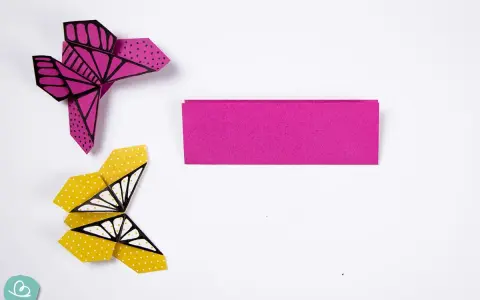 Zwei bunte origami schmetterlinge, einer pink mit Punkten, der andere gelb mit Punkten und Flügelmustern, liegen neben einem Streifen pinken Papiers.