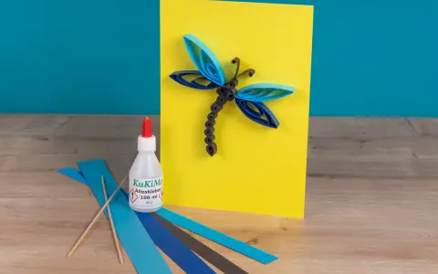 Eine Libelle aus Papierstreifen, erstellt mit einer Quilling-Anleitung. Kleber und bunte Streifen liegen bereit.