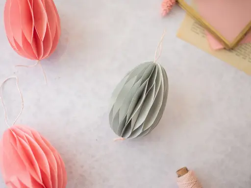 Ostereier basteln Papier: Dekorative Papier-Ostereier in Rosa und Grau hängen an Schnüren. Ein Garnknäuel und eine Schere liegen daneben.