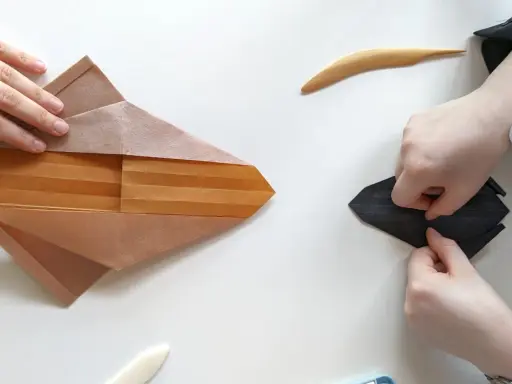 Zwei Hände falten Papier, eine an einem komplexen braunen Origami, die andere an einem schwarzen. Eine schwierige Origami-Anleitung wird befolgt.