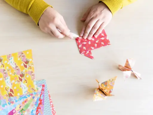 Hände falten Papier für eine Origami Kugel Anleitung. Mehrere bunte Origami-Papierbögen liegen bereit.