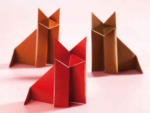Drei origami Füchse, einer rot und zwei braun, sitzen auf einem rosa Untergrund.