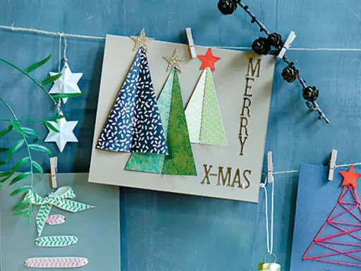 Selbstgebastelte Weihnachtskarten mit Bäumen und Sternen hängen an einer Schnur. Ein grüner Zweig mit Beeren und Papiersterne zieren die Szene.