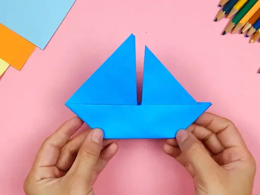 Hände falten ein blaues Papierboot. Bunte Stifte und Papier liegen bereit für weitere Bastelprojekte.