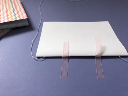 Beim Buch basteln werden weiße Blätter mit rosa Bändern und Faden zusammengefügt.