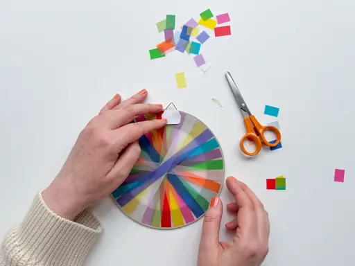 Hände basteln mit transparentsterne falten, bunte Streifen auf einem Kreis, daneben Schere und Konfetti.