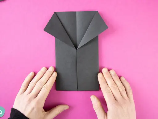 Ein schwarzes Origami-Hemd wird auf pinkem Hintergrund gefaltet. Hände halten das Papier.