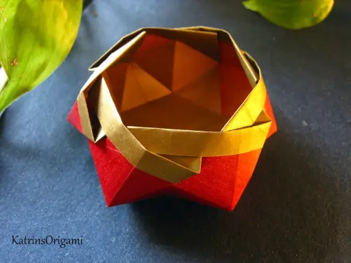 Eine kunstvolle origami schale in Rot und Gold, daneben ein grünes Blatt.