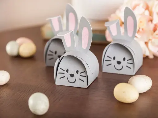 DIY Osternest: Süße Papierhasen mit rosa Ohren und Schokoladeneiern auf Holztisch.