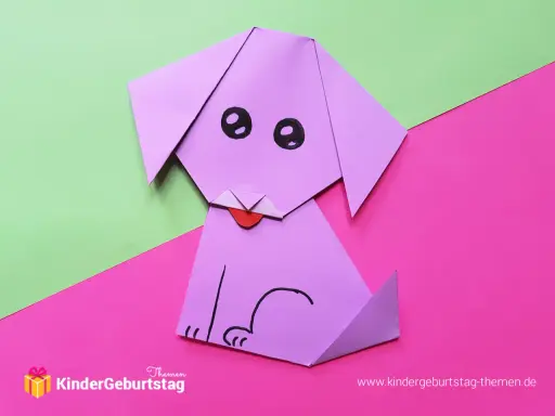Ein lila **Falten Hund** sitzt auf pinkem und grünem Papier. Er hat schwarze Augen und eine rote Zunge.