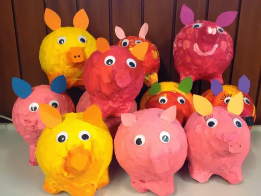 Bunte Pappmaché-Sparschweine mit großen Kulleraugen und Papierohren stehen fröhlich nebeneinander.
