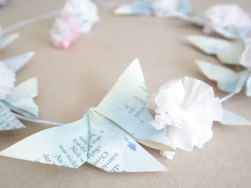 Zarte Origami-Schmetterlinge aus Buchseiten, verziert mit Tupfen und kleinen Papierblumen, bilden einen Kranz. Ein Hauch von origami frühling.