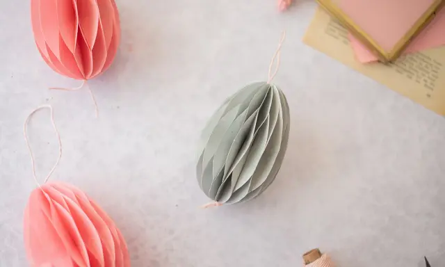 Ostereier basteln Papier: Dekorative Papier-Ostereier in Rosa und Grau hängen an Schnüren. Ein Garnknäuel und eine Schere liegen daneben.
