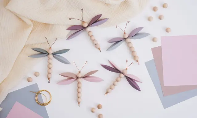 Vier selbstgebastelte Libellen aus Holzperlen und Papierflügeln, inspiriert von einer libelle basteln vorlage. Zarte Pastellfarben und ein Musselintuch schaffen eine frühlingshafte Atmosphäre.
