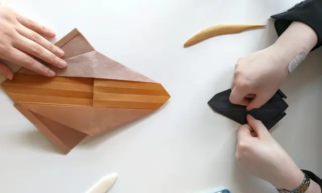 Zwei Hände falten Papier, eine an einem komplexen braunen Origami, die andere an einem schwarzen. Eine schwierige Origami-Anleitung wird befolgt.