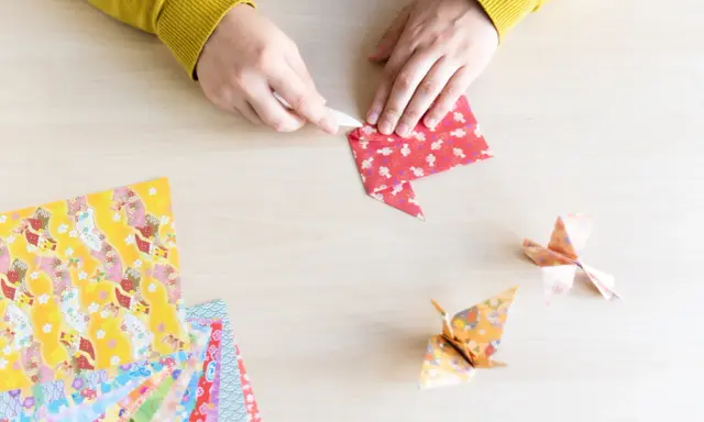 Hände falten Papier für eine Origami Kugel Anleitung. Mehrere bunte Origami-Papierbögen liegen bereit.