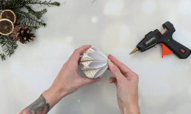 Hände basteln mit Klebepistole und Papier einen schönen DIY Christbaumschmuck. Tannenzweige und Orangenscheiben liegen bereit.