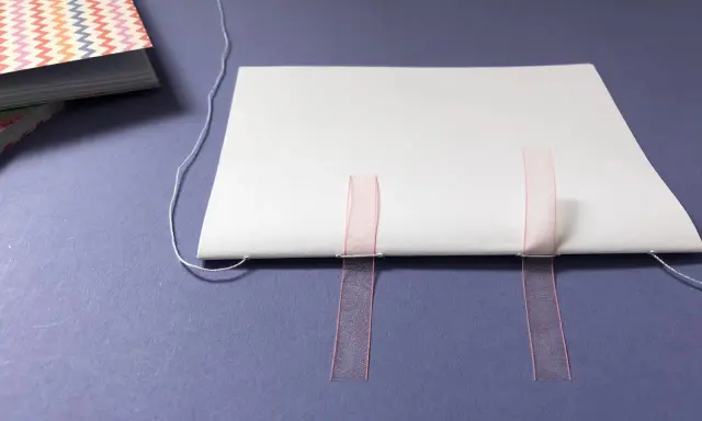 Beim Buch basteln werden weiße Blätter mit rosa Bändern und Faden zusammengefügt.