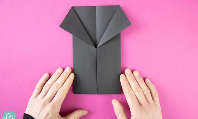 Ein schwarzes Origami-Hemd wird auf pinkem Hintergrund gefaltet. Hände halten das Papier.