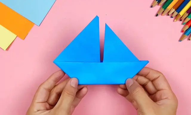 Hände falten ein blaues papierschiffchen. Bunte Stifte und Papier liegen bereit, um das papierschiffchen bauen zu beginnen.