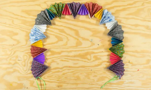 Bunte Papierfächer, die zu einer ziehharmonika-girlande gebastelt wurden, liegen auf einer Holzplatte.