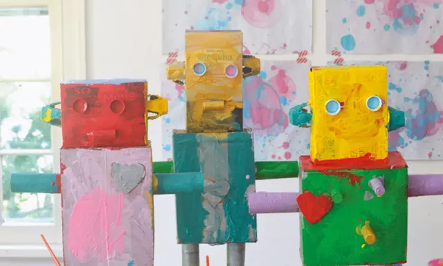 Drei bunte Roboter, die Kinder gebastelt haben, stehen nebeneinander. Sie sind aus Kartons und Rollen gefertigt und mit leuchtenden Farben bemalt.