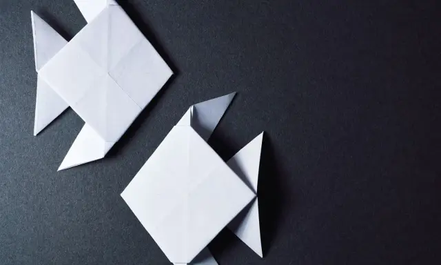 Zwei weiße Origami-Fische, kunstvoll gefaltet, schwimmen auf dunklem Grund.