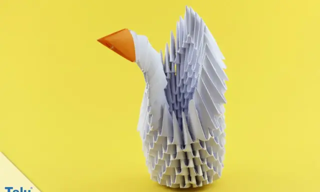 Ein weißer Schwan aus modularem Origami mit orangefarbenem Schnabel steht vor einem gelben Hintergrund.