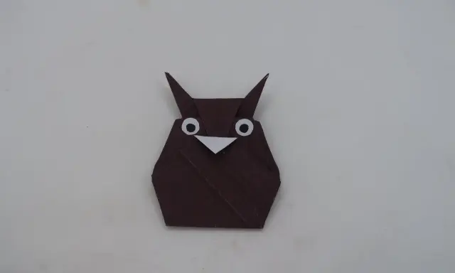 Eine braune origami Eule mit großen weißen Augen und einem kleinen Schnabel sitzt auf hellem Untergrund.